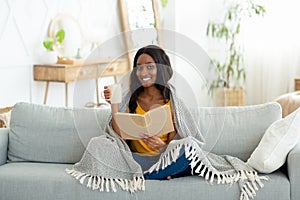 Lovely black woman reading book, covered with warm plaid, drinking hot aromatic coffee on sofa at home