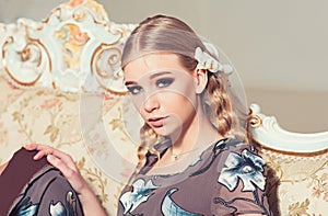 Lovely blond girl with curly hair sitting on vintage sofa. Closeup portrait of young lady in flowery dress