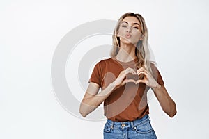 Lovely blond girl pucker lips in kiss, showing heart love gesture on chest, standing over white background