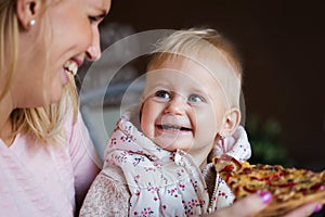 Lovely blond little baby girl in beautiful white dress biting on piece of tasty pizza