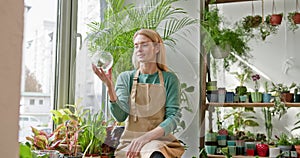 Lovely blond middle age female owner of plant store with flower in pot and gardening set in shop interior. A female