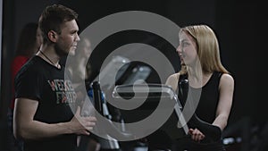 Lovely blonde girl is diligent working on an exercise bike and cool guy come to say hi on a background of practiced