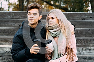 Lovely brunette guy and pretty girl blonde drink coffee and chat on date, dreaming. Loving teenagers are sitting on park bench.