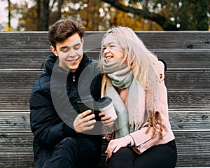 Lovely brunette guy and pretty girl blonde drink coffee and chat on date, laugh. Loving teenagers are sitting on park bench. Teen