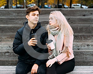 Lovely brunette guy and pretty girl blonde drink coffee and chat on date. Loving teenagers are sitting on park bench. Teen Love