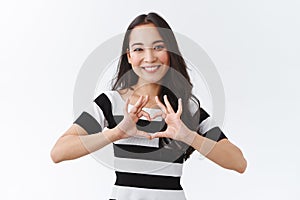 Lovely, caring and outgoing east-asian woman in striped t-shirt, showing heart sign over chest and smiling with tender