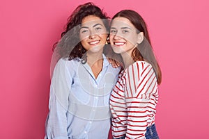 Lovely Caucasian females embracing and smiling to camera, posing isolated over pink background, dresses stylish shirts,