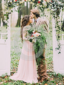 Lovely close-up portrait of the newlyweds standing under the white arch in the form of two doors decorated with the red