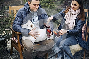 Lovely couple with cute dog sitting on backyard outside surrounded by young coniferous trees
