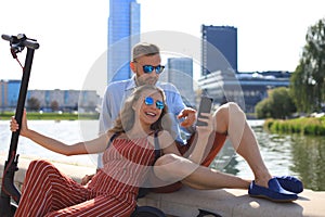 Lovely couple having fun driving electric scooter, taking a break from driving, sitting on riverbank, taking selfie