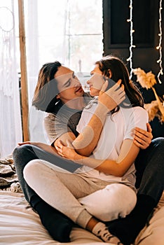 Lovely couple hugging on their bed decorated with garland lights at home indoors in the bedroom