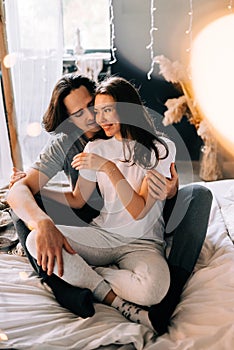 Lovely couple hugging on their bed decorated with garland lights at home indoors in the bedroom