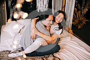 Lovely couple hugging on their bed decorated with garland lights at home indoors in the bedroom