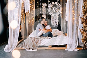 Lovely couple hugging on their bed decorated with garland lights at home indoors in the bedroom