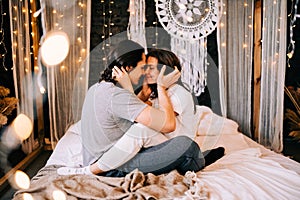 Lovely couple hugging on their bed decorated with garland lights at home indoors in the bedroom