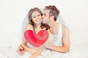 Lovely couple in love holding red paper heart in bed and kissing
