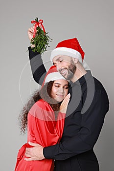 Lovely couple in Santa hats under mistletoe bunch on grey background