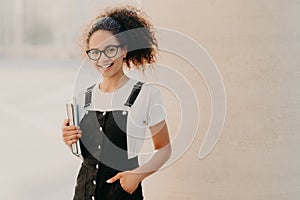 Lovely curly woman with combed hair, dressed in white t shirt, sarafan, keeps hand in pocket, holds book and textbook, wears