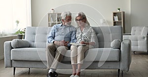 Lovely elderly couple reading book sitting on sofa at home