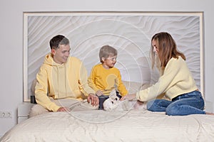 Lovely family, cute parents, mom and dad with adorable child son sitting on the bed and playing with little white dog