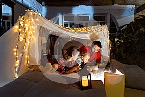 Lovely family, mother, father and daughter lying inside self-made hut, tent in room in the evening with Christmas lights