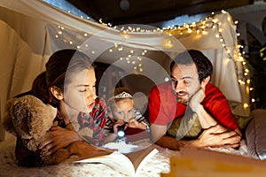 Lovely family, mother, father and daughter lying inside self-made hut, tent in room in the evening and reading book