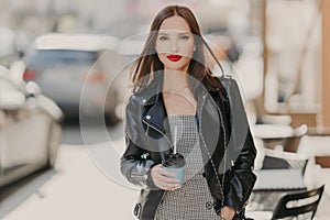 Lovely fashionable woman with makeup, bright red painted lips, wears black leather jacket, strolls at urban setting, stands near