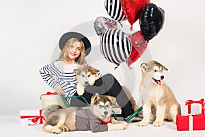 Lovely girl with blond hair wearing a hat with Malamute puppies with bright balloons hearts and gifts for Valentine`s Day