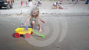 Lovely girl with curly blond hair digging sand to toy cart wagon on ocean shore