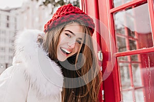 Lovely girl with happy smile posing close to red phone booth in december morning. Outdoor portrait of wonderful european