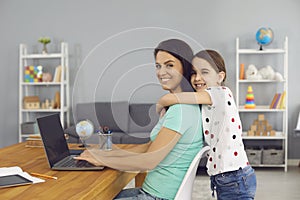 Lovely girl hugging her mom while she working online via laptop at home. Cute child embracing her mother indoors