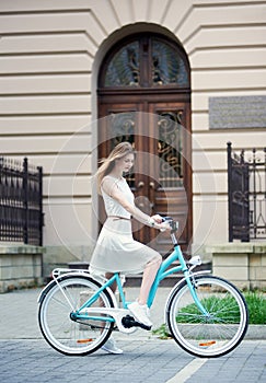 Lovely girl in light dress and sneakers on blue retro bike against the background of an old building
