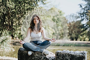 Lovely girl meditating alone in the park, sitting on a big rock near beautiful small lake.