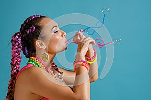 Lovely girl with a multi-colored braids hairstyle and bright make-up, is blowing bubbles using tubules, posing in studio