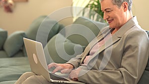 Lovely grey-haired grandmother with a short haircut wearing a gray business suit works sitting on the sofa in the living