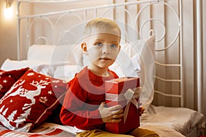 Lovely happy child boy in red sweater sitting on white bed with red pillows holding Christmas gift in hands. Christmas concept