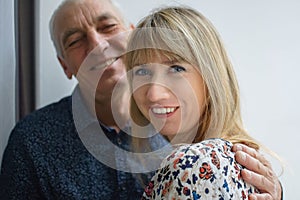 Lovely happy portrait of elderly man hugging his young blonde-haired smiling wife wearing warm dress. Couple with age