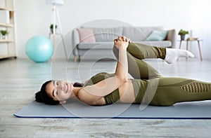 Lovely Indian woman stretching her leg while lying on mat indoors. Stay home sports