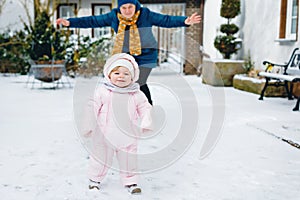 Lovely little baby girl making first steps outdoors in winter. Cute toddler learning walking. Happy mother on background