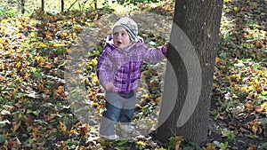 Lovely little baby girl standing near tree trunk and falling autumn leaves. 4K