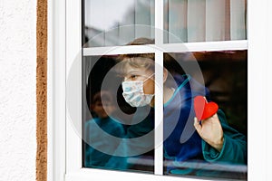 Lovely little school kid boy by a window wearing medical mask and holding wooden heart during pandemic coronavirus