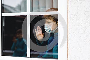 Lovely little school kid boy by a window wearing medical mask at home or school during pandemic coronavirus quarantine