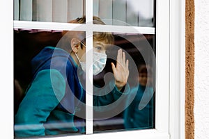 Lovely little school kid boy by a window wearing medical mask at home or school during pandemic coronavirus quarantine