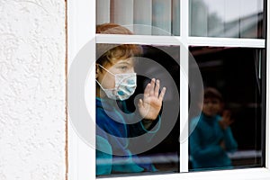 Lovely little school kid boy by a window wearing medical mask at home or school during pandemic coronavirus quarantine