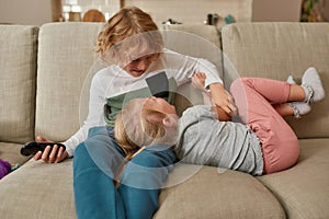 Lovely little siblings, boy and girl having fun, playing together while watching TV, cuddling on a sofa at home