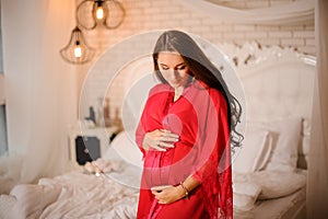 Lovely long-haired pregnant woman dressed in elegant pink negligee standing near the bed