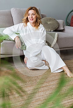 Lovely middle-aged blond woman with a beaming smile sitting on the floor close to the sofa