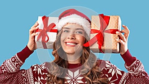 Lovely millennial lady with two Christmas gifts wearing Santa hat and smiling on blue bckground