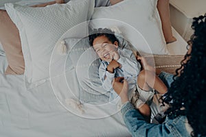 Lovely mother tickling her cute little son lying on bed and laughing