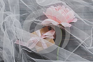 Lovely pink peony flower and gift box under soft tulle fabric on dark wood, top view. Mothers day